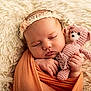 baby, sleeping, swaddled, orange_wrap, pink_teddy_bear, crocheted_toy, headband, lace, blanket, fluffy, soft_texture, infant, cute, peaceful, portrait, closeup, hand, face, resting, indoor