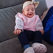 Elya participe au concours pour gagner de l'argent avec cette photo : baby, blue_socks, child, clothing, couch, cozy, cute, fabric, footwear, happy, headband, home, indoor, infant, person, pink_sweater, portrait, seated, smiling, white_shoes