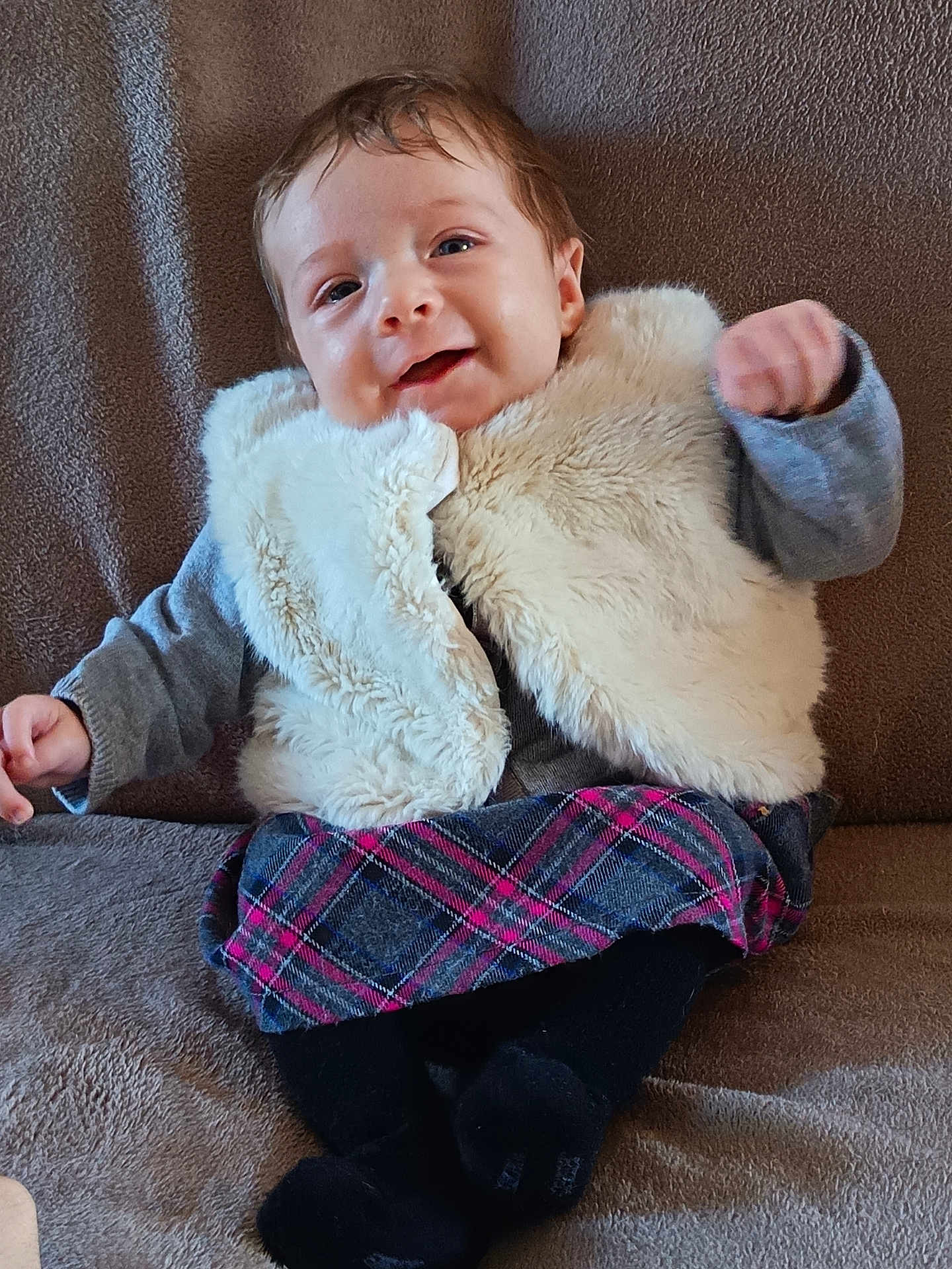 Elya a rejoint le concours — aidez-le/la à gagner de superbes lots ! baby, child, smiling, sitting, vest, plaid_skirt, black_tights, couch, brown_background, happy, fuzzy_clothing, indoor, portrait, cute, infant, young_child, furniture, person, cozy, raised_fist