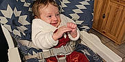 Elya participe au concours pour gagner de l'argent avec cette photo : baby, child, high_chair, smiling, cozy_clothing, indoor, kitchen, wooden_cabinet, stone_wall, cushion, seat_belt, floor_tile, happy, person, infant, sitting, warm_lighting, red_pants, white_jacket, sock