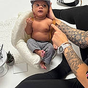 Dominic is registered to the contest to win money with this photo: baby, infant, hat, pants, knit_chair, tattoos, watch, camera, checkered_strap, pointing_hand, adult, bare_chest, curious_expression, white_background, plant, table, black_clothing, socks, bracelet, indoor