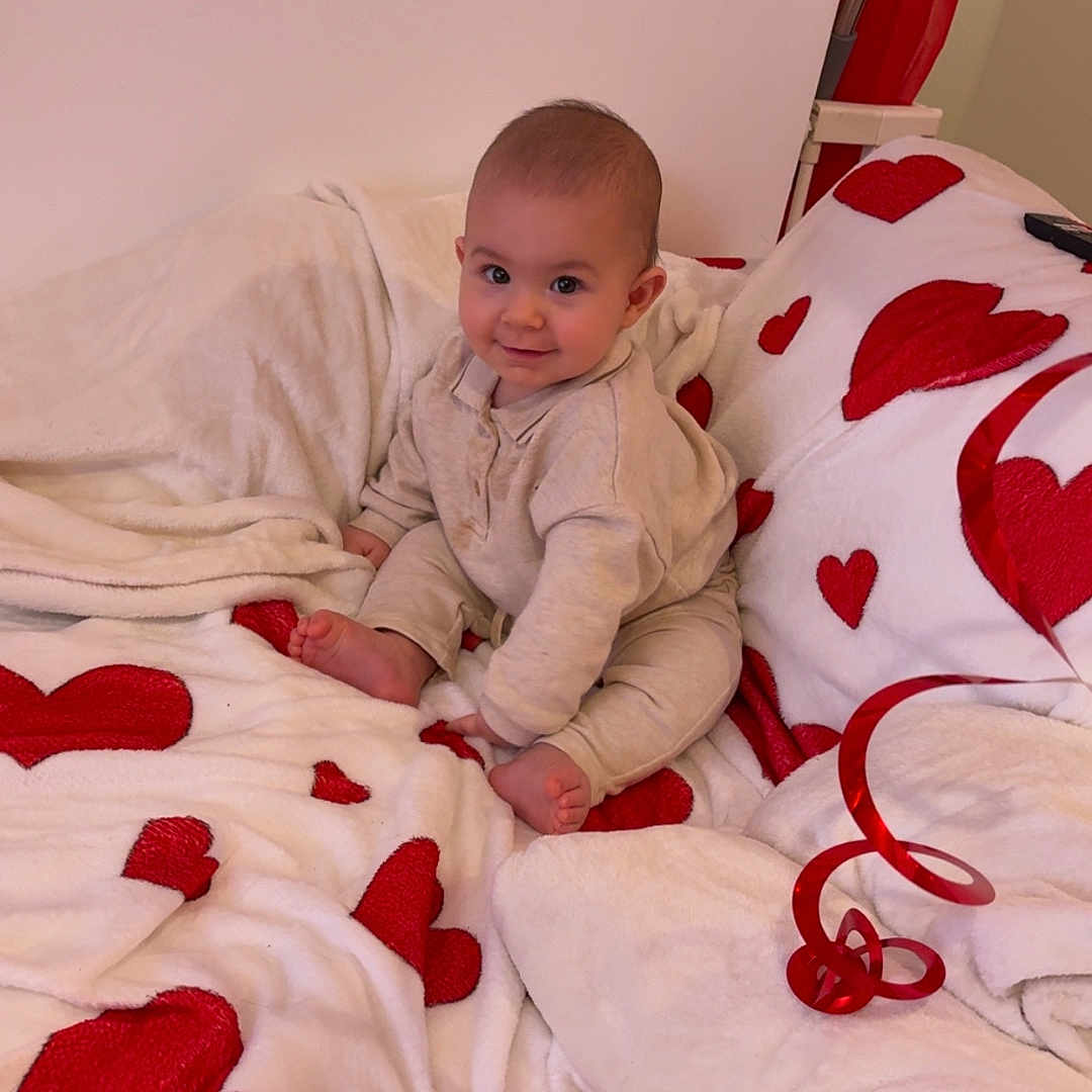 Jules a rejoint le concours — aidez-le/la à gagner de superbes lots ! baby, child, smile, blanket, red_hearts, cozy, home_interior, cute, infant, sitting, soft_toys, floor, furniture, warm_colors, happy, portrait, candid, cute_baby, indoor, plush