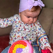 Andréa a rejoint le concours — aidez-le/la à gagner de superbes lots ! baby, bow, child, closeup, colorful, couch, cute, denim, floral_clothing, headband, indoor, infant, jeans, person, playing, pop_it_toy, rainbow_colors, red_toy, soft_lighting, toy