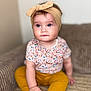 baby, blue_eyes, bow, child, couch, cozy, cute, floral_shirt, foot, hand, headband, indoors, infant, person, portrait, sitting, skin, soft_background, yellow_pants, young