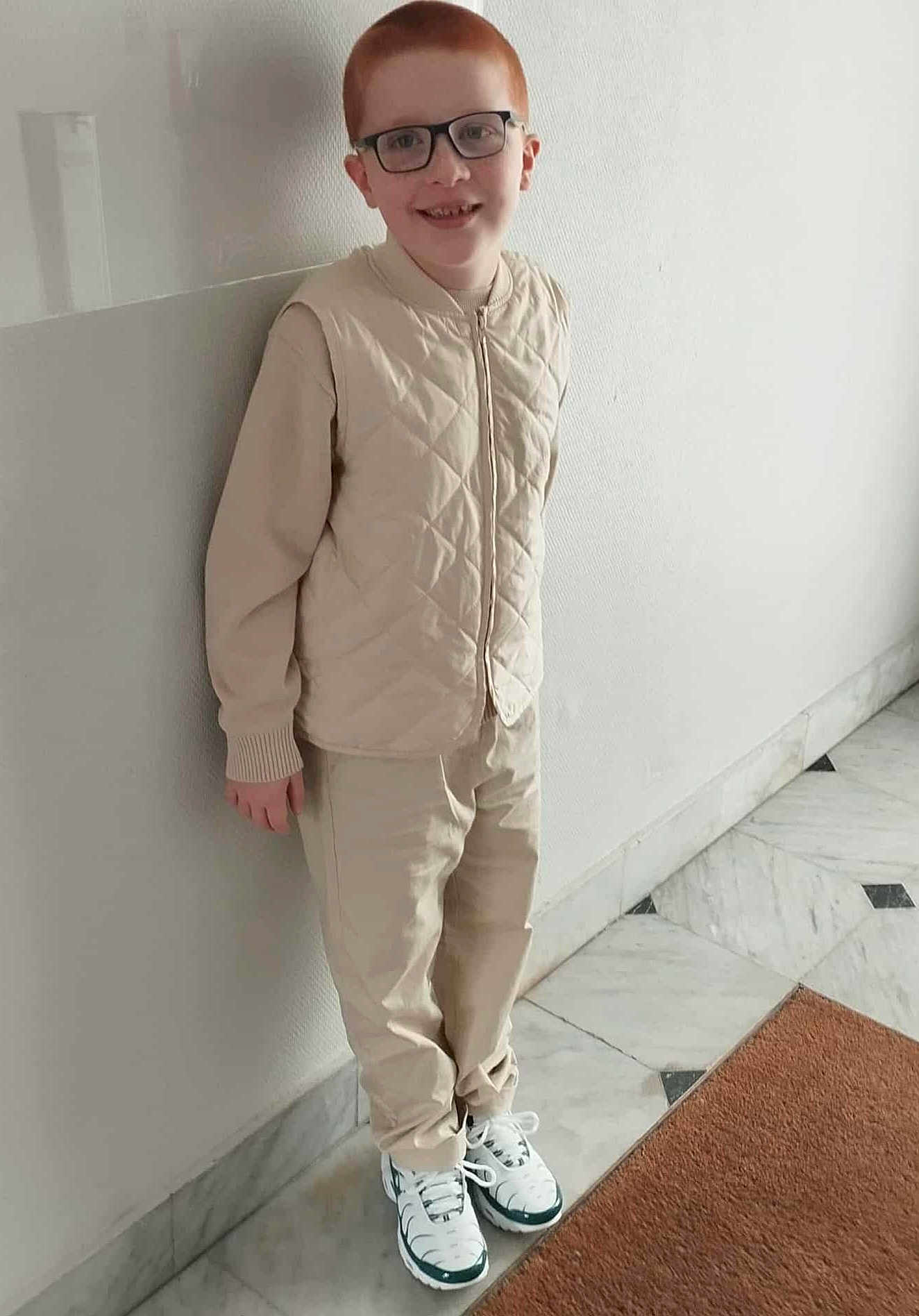 Kyllian a rejoint le concours — aidez-le/la à gagner de superbes lots ! child, red_hair, glasses, smiling, standing, beige_clothing, jacket, pants, sneakers, marble_floor, doormat, indoor, wall, person, casual, portrait, happy, young, fashion, feet