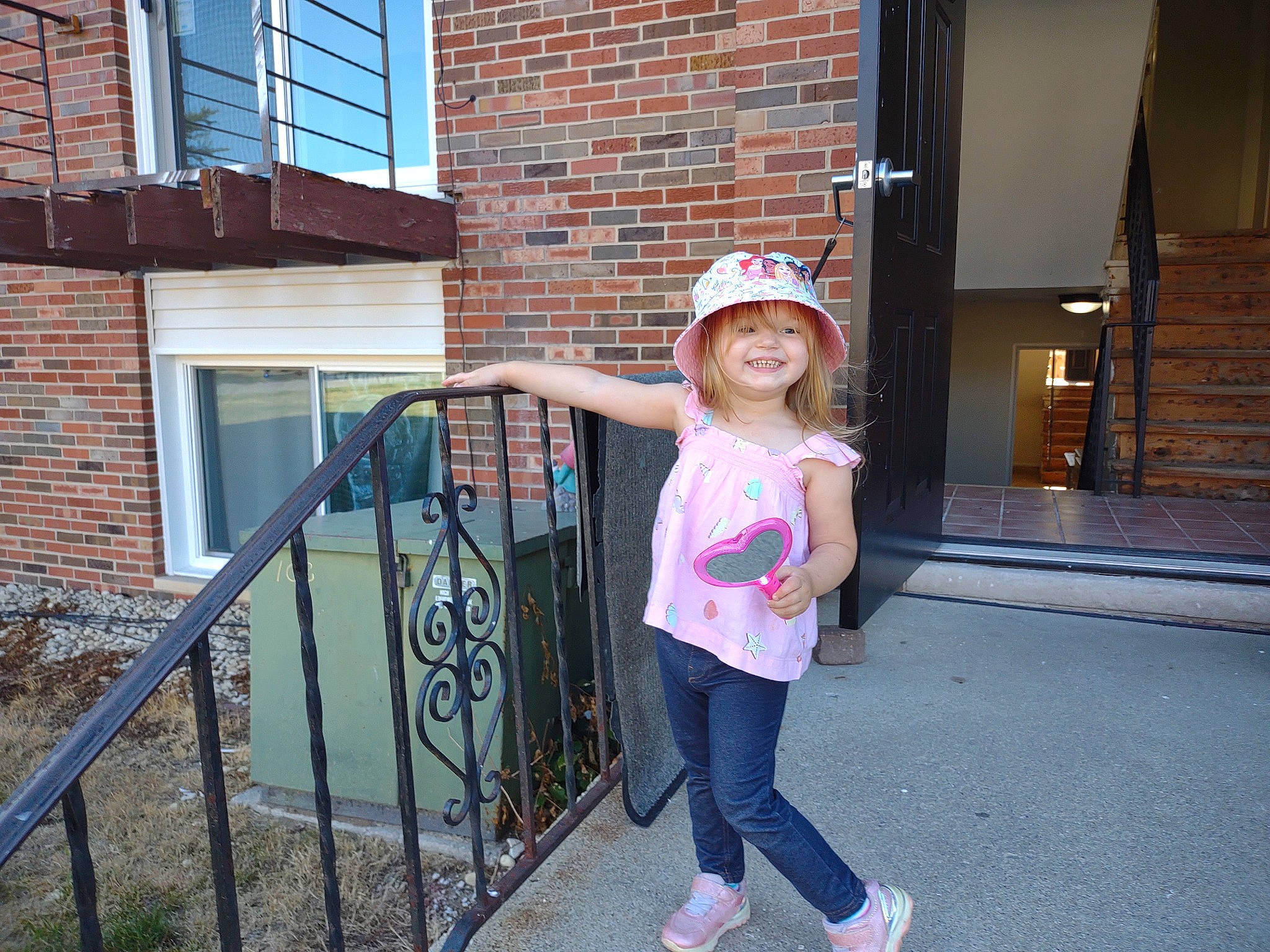 Abigail is registered to the contest to win money with this photo: blue, brick, brickwork, building, cap, electric_blue, happy, hat, headwear, house, joy, leisure, person, sleeve, smile, standing, sun_hat, t_shirt, toddler, tree