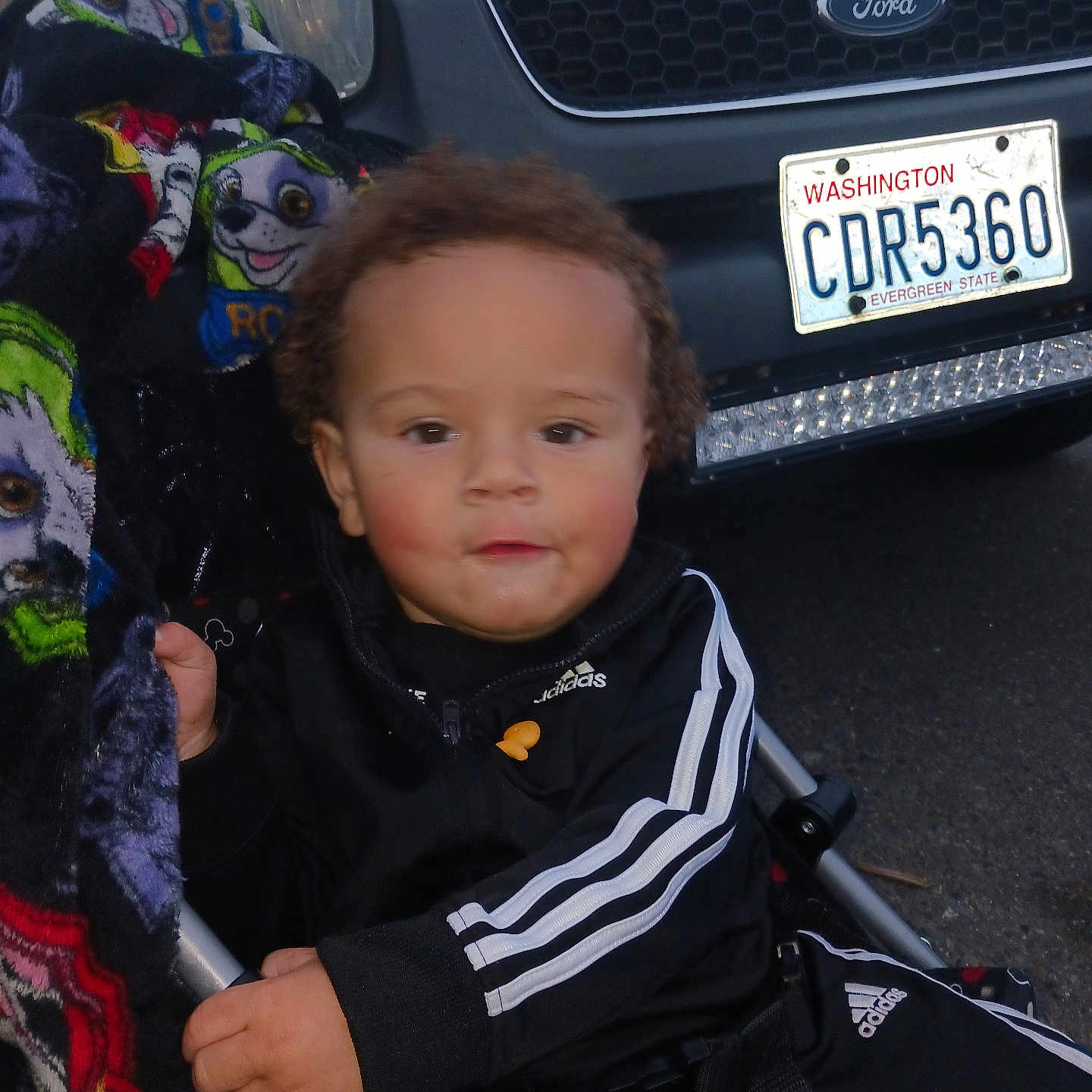 Loki is registered to the contest to win money with this photo: baby, child, infant, portrait, sitting, stroller, automobile, ford, license_plate, outdoor, blanket, colorful, tracksuit, adidas, brown_hair, face, eyes, rosy_cheeks, vehicle, photography