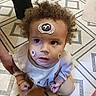 baby, child, curly_hair, sticker, spider, face, portrait, looking_up, indoor, tile_floor, patterned_floor, infant, clothing, shirt, pants, adult_hand, sitting, curious, closeup, photography