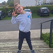 Vicenzo participe au concours pour gagner de l'argent avec cette photo : child, boy, glasses, smiling, outdoor, steps, casual_clothing, sneakers, hand_gesture, building, car, parking_lot, hedge, overcast_sky, sidewalk, railing, grass, playful, young_person, urban