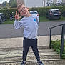 child, boy, glasses, smiling, outdoor, steps, casual_clothing, sneakers, hand_gesture, building, car, parking_lot, hedge, overcast_sky, sidewalk, railing, grass, playful, young_person, urban