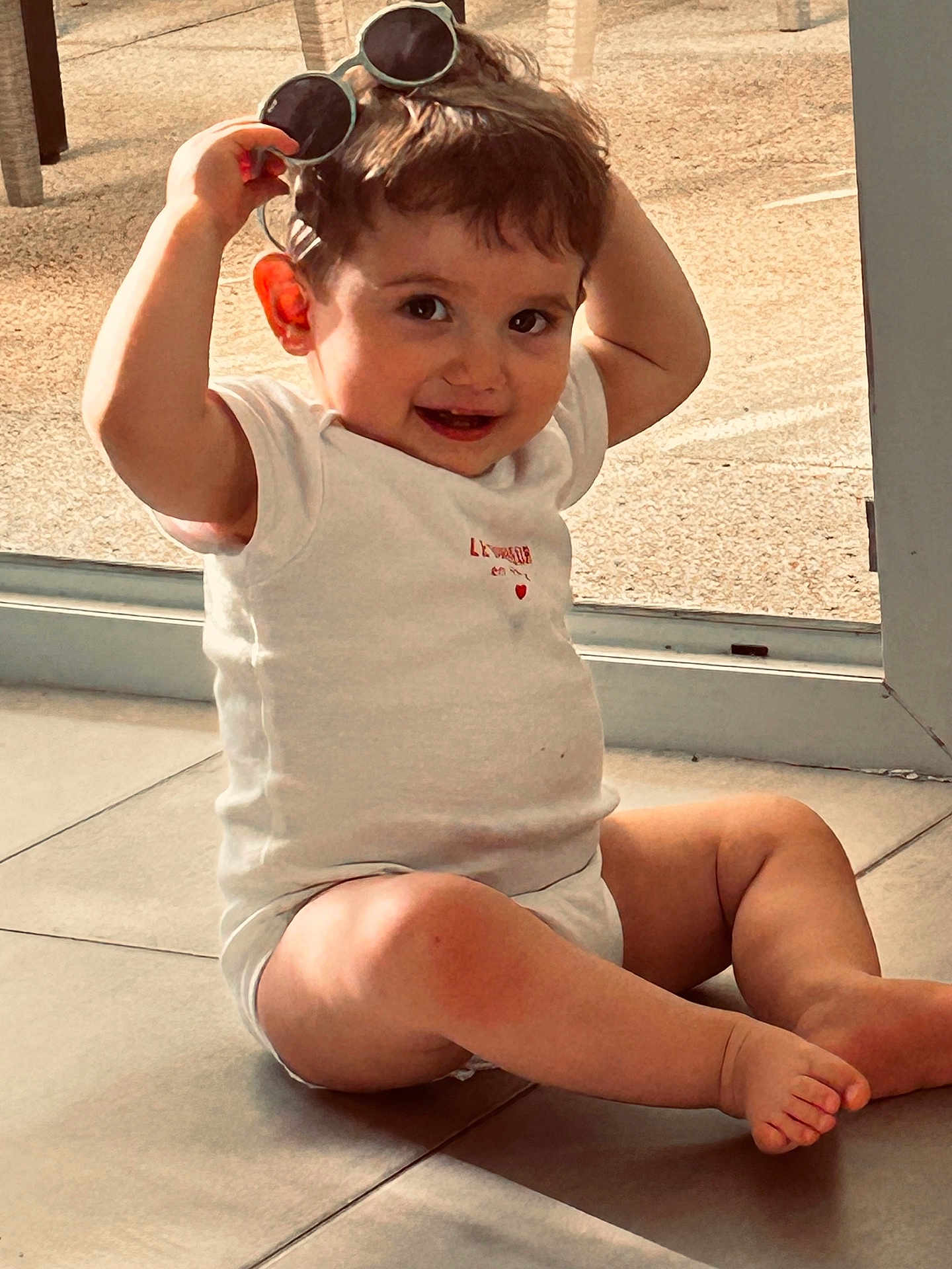 Lino participe au concours pour gagner de l'argent avec cette photo : toddler, child, indoor, floor, sunglasses, smile, playful, white_clothing, barefoot, window, natural_light, happy, cute, baby, person, young_child, skin, legs, head, expression