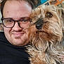 man, dog, glasses, selfie, smile, indoor, portrait, furry_dog, brown_dog, casual_clothing, face, wall_decor, photographs, home, cuddling, closeup, happy, pet, friendship, cozy