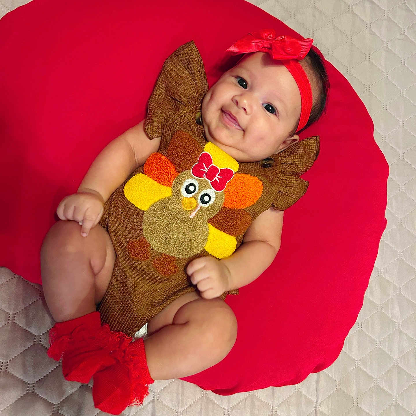 Rachell is registered to the contest to win money with this photo: apparel, baby, brown_outfit, child, cushion, cute, happy, headband, indoors, infant, lying_down, person, plush, quilted_bedspread, red_cushion, red_headband, red_socks, smiling, sock, turkey_design
