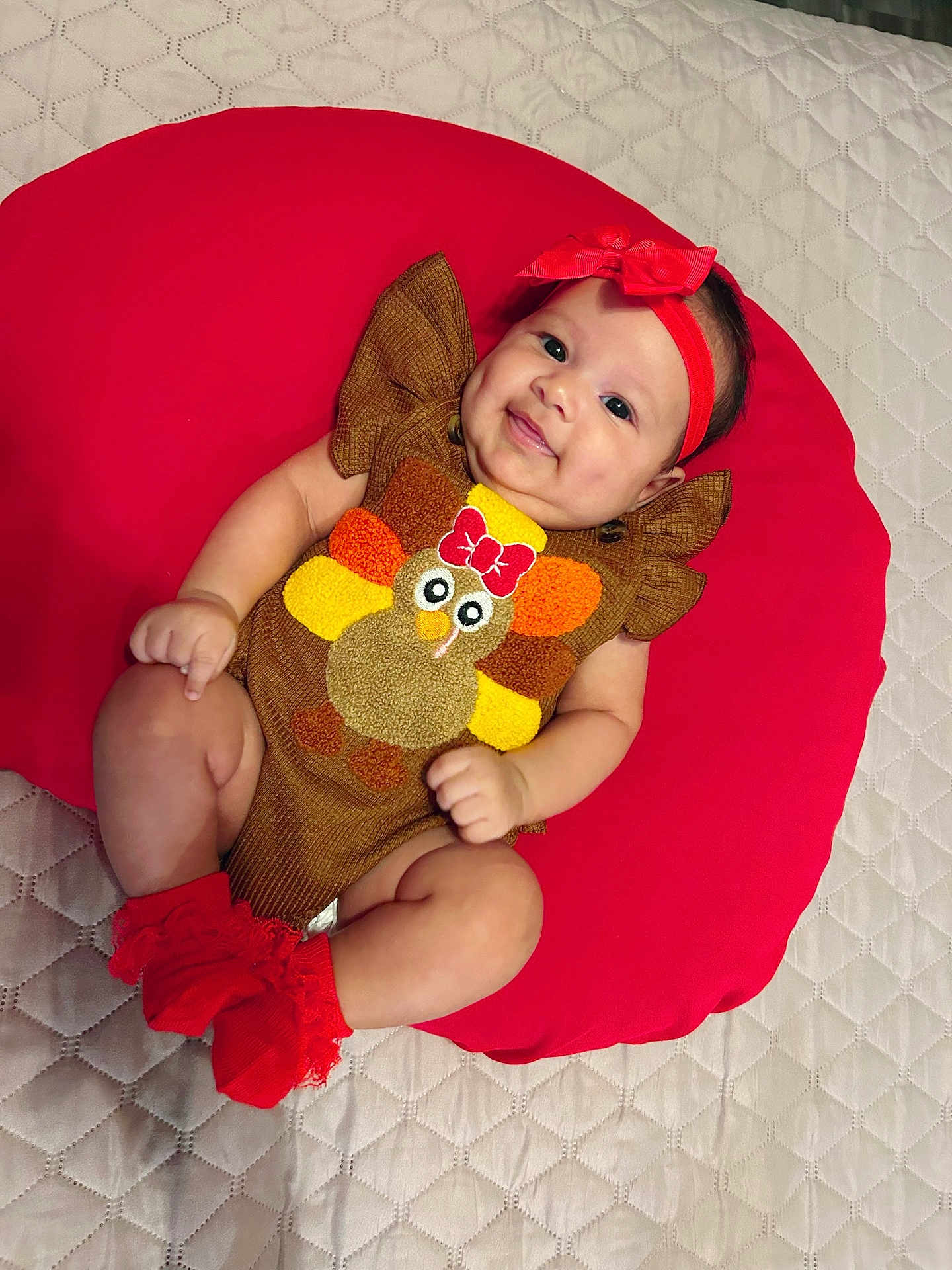 Rachell is registered to the contest to win money with this photo: baby, infant, smiling, red_headband, red_socks, brown_outfit, turkey_design, cushion, red_cushion, quilted_bedspread, cute, happy, lying_down, indoors, child, person, headband, sock, plush, apparel