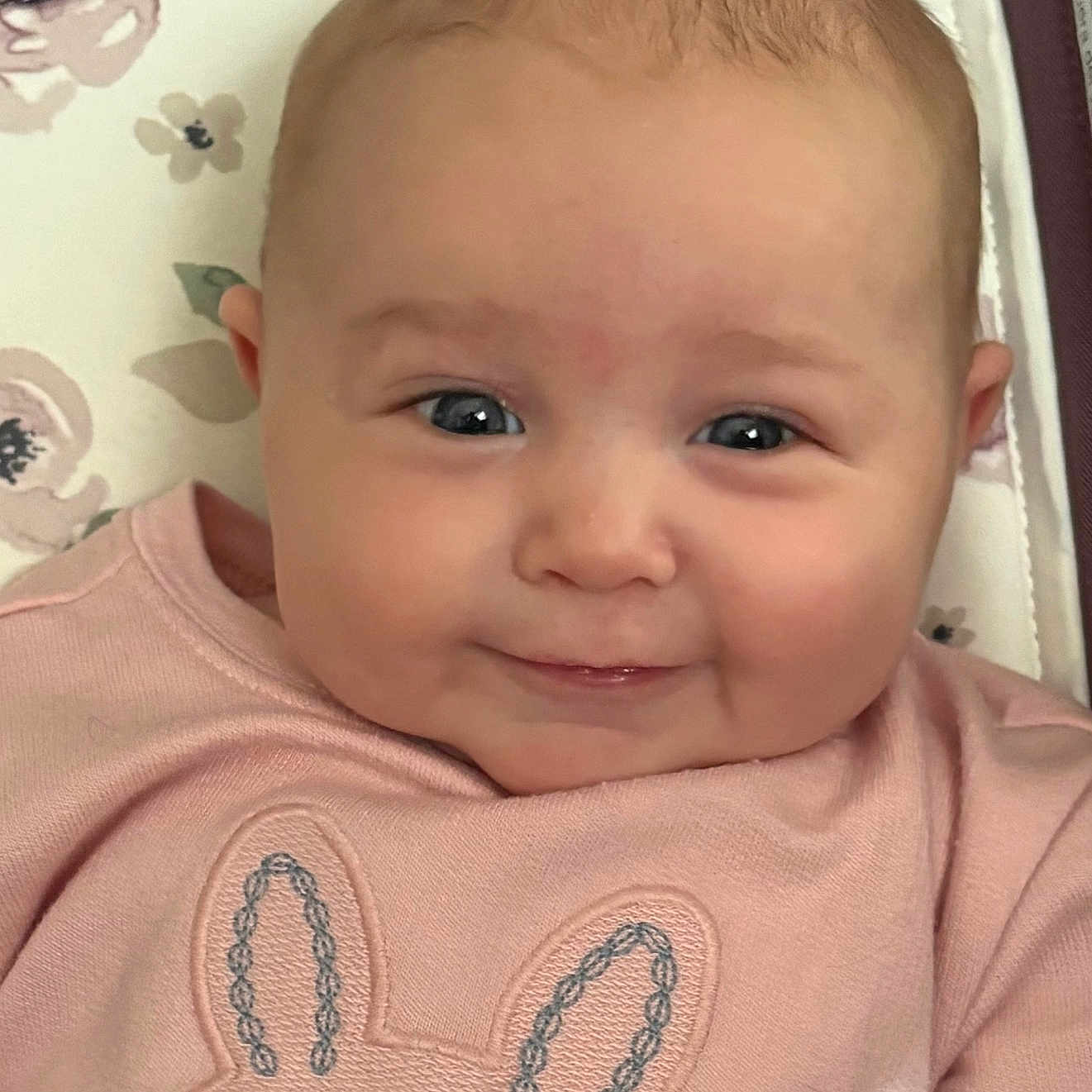 Maura joined the competition — help win amazing prizes! baby, infant, smile, face, pink_clothing, bunny_design, cushion, floral_pattern, cute, child, portrait, closeup, happy, indoors, person, head, cheeks, eyes, baby_girl, soft_lighting