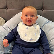Bowen is registered to the contest to win money with this photo: baby, bed, bib, blanket, blue_clothing, child, cozy, cute, face, happy, headboard, indoors, infant, person, pillow, portrait, sitting, smiling, sock, toddler