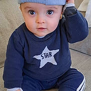 Liam a rejoint le concours — aidez-le/la à gagner de superbes lots ! baby, child, blue_cap, sweater, star, barefoot, couch, indoor, curious, sitting, casual_clothing, infant, portrait, cute, hand_on_cap, young_child, relaxed, cozy, smiling, person