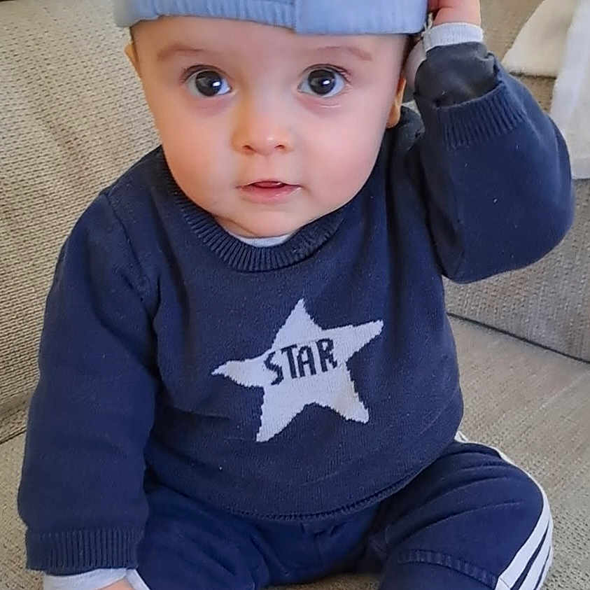 Liam a rejoint le concours — aidez-le/la à gagner de superbes lots ! baby, child, blue_cap, sweater, star, barefoot, couch, indoor, curious, sitting, casual_clothing, infant, portrait, cute, hand_on_cap, young_child, relaxed, cozy, smiling, person