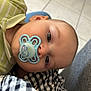 baby, pacifier, green_shirt, striped_clothing, infant, child, tiled_floor, lap, closeup, person, face, head, skin, hand, sitting, indoor, young_child, clothing, comfort, rest