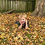 child, boy, smiling, autumn, leaves, grass, tree, fence, outdoor, fall, camo_shirt, boots, nature, playful, portrait, casual, seasonal, young, happy, daylight