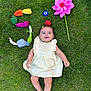 baby, child, grass, outdoor, dress, plush_toys, vegetables, pinwheel, greenery, smiling, barefoot, playful, toy, summer, happy, nature, lying_down, colorful, cute, fun