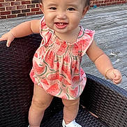 Aliyah is registered to the contest to win money with this photo: toddler, child, smiling, standing, wicker_chair, wooden_deck, brick_wall, window, dress, watermelon_pattern, white_shoes, earring, bracelet, outdoor, happy, cute, person, face, baby, summer