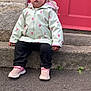 casual_clothing, casual_pose, child, curious_expression, curly_hair, cute, daylight, outdoor, person, pink_shoes, portrait, raincoat, red_door, sidewalk, sitting, small_plant, stone_step, street, toddler, young_child