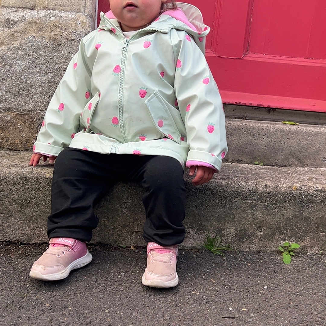 Nairobi-Louise is registered to the contest to win money with this photo: casual_clothing, casual_pose, child, curious_expression, curly_hair, cute, daylight, outdoor, person, pink_shoes, portrait, raincoat, red_door, sidewalk, sitting, small_plant, stone_step, street, toddler, young_child