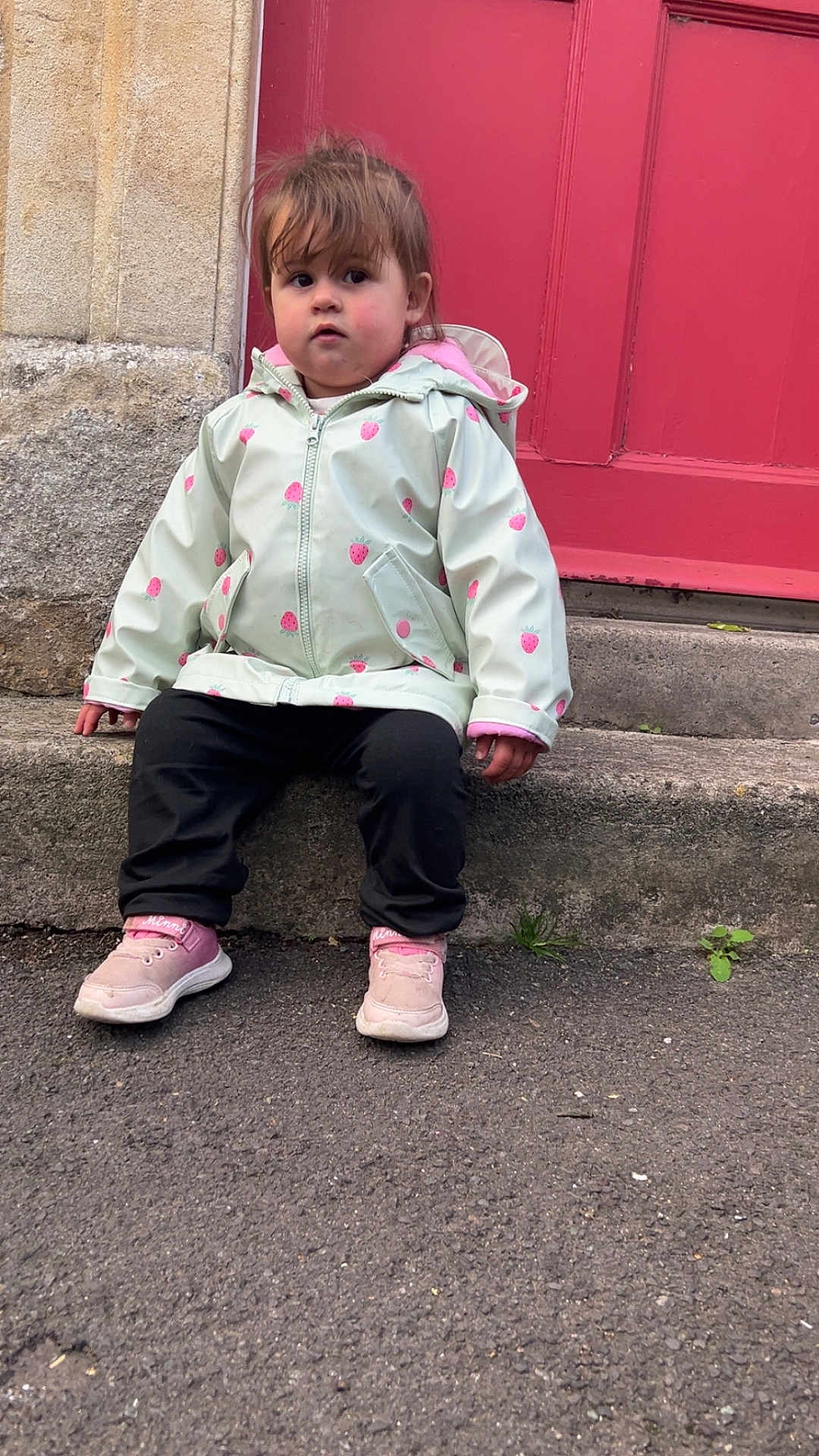 Nairobi-Louise is registered to the contest to win money with this photo: casual_clothing, casual_pose, child, curious_expression, curly_hair, cute, daylight, outdoor, person, pink_shoes, portrait, raincoat, red_door, sidewalk, sitting, small_plant, stone_step, street, toddler, young_child