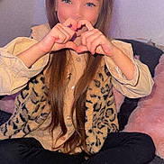 Myla a rejoint le concours — aidez-le/la à gagner de superbes lots ! child, girl, hand_gesture, heart_shape, long_hair, bow, leopard_print, vest, beige_shirt, black_leggings, barefoot, rug, cushions, indoor, smiling, seated, cozy, portrait, happy, casual
