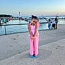 child, girl, pink_outfit, playful_pose, smiling, marina, waterfront, lighthouse, boats, yacht, sailboat, harbor, dock, seaside, outdoor, sunset, travel, portrait, fashion, blue_shoes