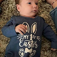 Michael is registered to the contest to win money with this photo: baby, infant, onesie, blue_clothing, carpet, floor, feet, shoes, person, child, laying_down, looking_away, indoor, cute, small_hand, text_on_clothing, celebration, easter, casual, relaxed