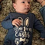 baby, infant, onesie, blue_clothing, carpet, floor, feet, shoes, person, child, laying_down, looking_away, indoor, cute, small_hand, text_on_clothing, celebration, easter, casual, relaxed