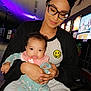 woman, baby, glasses, headband, hair_buns, smile, holding, arcade, colorful_lights, indoor, casual_clothing, baby_pajamas, floral_pattern, seated, portrait, happy, person, technology, neon_lights, family