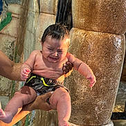 Leandro C is registered to the contest to win money with this photo: baby, child, crying, water, waterfall, wet_hair, swim_trunks, adult_hands, holding, stone_wall, splash, pool, waterpark, tears, wet_skin, shorts, expression_crying, indoor, grip, toddler
