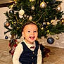 child, toddler, smiling, christmas_tree, ornaments, lights, holiday, indoor, floor, barefoot, vest, bow_tie, happy, celebration, festive, decorations, person, clothing, young_child, seated