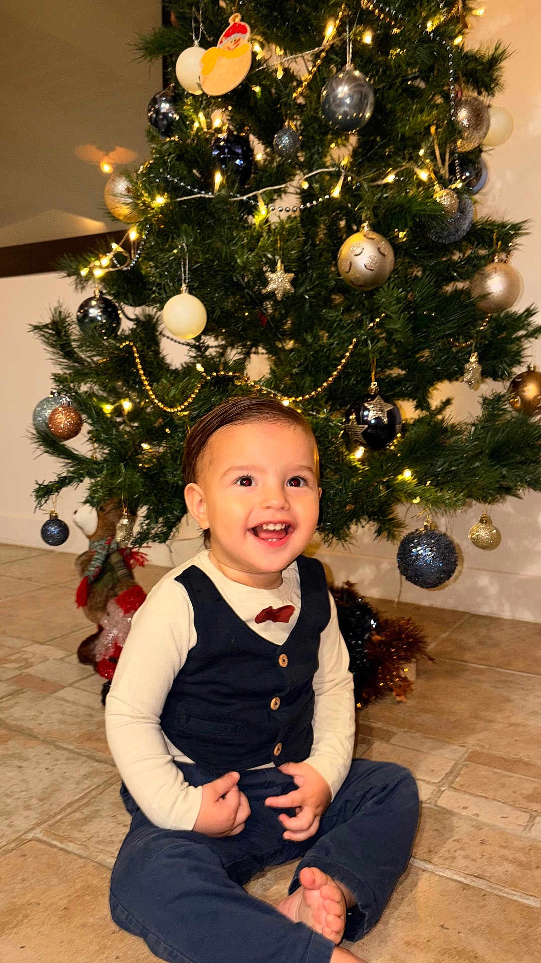 Milaan participe au concours pour gagner de l'argent avec cette photo : child, toddler, smiling, christmas_tree, ornaments, lights, holiday, indoor, floor, barefoot, vest, bow_tie, happy, celebration, festive, decorations, person, clothing, young_child, seated