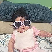 Kaylee is registered to the contest to win money with this photo: baby, infant, child, portrait, sunglasses, heart_shaped_sunglasses, pink_clothing, pajamas, sitting, bed, blanket, bedroom, indoor, patterned_bedding, dark_hair, face, fashion, cute, casual, photography