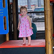Skylar is registered to the contest to win money with this photo: toddler, child, pink_dress, barefoot, playground, play_structure, orange_platform, mesh_screen, wooden_beam, smile, standing, outdoor, daylight, curly_hair, cute, patterned_clothing, vehicle, window, person, expression