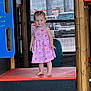 toddler, child, pink_dress, barefoot, playground, play_structure, orange_platform, mesh_screen, wooden_beam, smile, standing, outdoor, daylight, curly_hair, cute, patterned_clothing, vehicle, window, person, expression