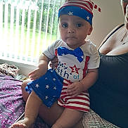 Brandon is registered to the contest to win money with this photo: baby, infant, child, person, indoor, window, clothing, hat, stars, stripes, red, white, blue, sitting, adult, hand, bed, blanket, curious, portrait