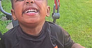 Solomon is registered to the contest to win money with this photo: child, smiling, outdoor, sunlight, grass, stroller, black_shirt, happy, portrait, joyful, casual_clothing, person, daylight, nature, playful, young, cute, fun, hands, sitting