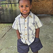 Solomon is registered to the contest to win money with this photo: child, boy, smiling, standing, plaid_shirt, shorts, sneakers, concrete_floor, refrigerator, wooden_panel, outdoor, happy, casual_clothing, portrait, hands_in_pockets, curly_hair, daylight, person, young, confident