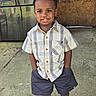 child, boy, smiling, standing, plaid_shirt, shorts, sneakers, concrete_floor, refrigerator, wooden_panel, outdoor, happy, casual_clothing, portrait, hands_in_pockets, curly_hair, daylight, person, young, confident