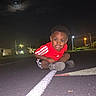 Solomon is registered to the contest to win money with this photo: child, night, running_track, red_shirt, sneakers, flash, moon, clouds, outdoor, street_lights, playful, crouching, sporty, young_boy, dark_sky, asphalt, line_marking, urban, expression, casual_clothing