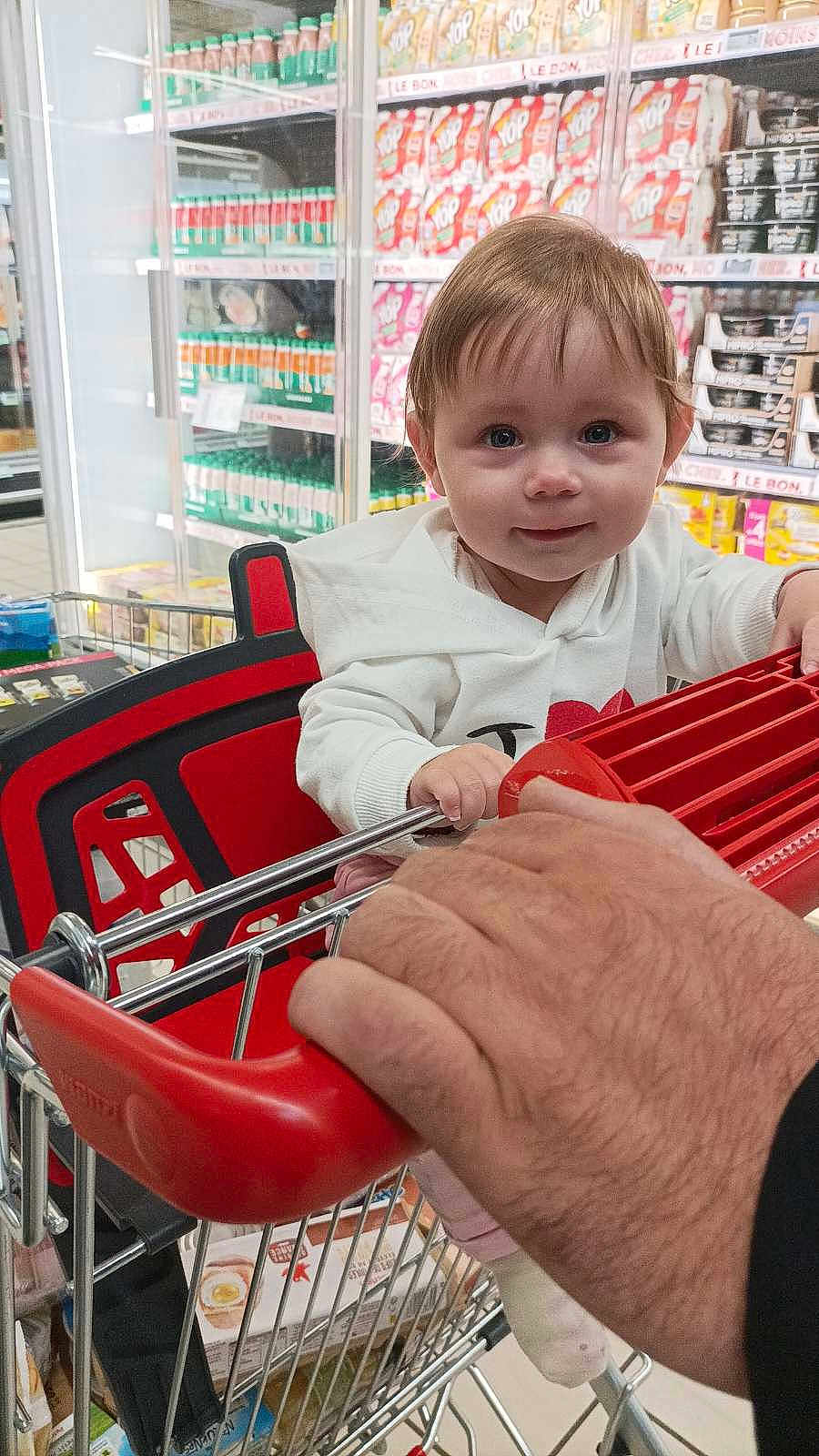 Lea a rejoint le concours — aidez-le/la à gagner de superbes lots ! baby, child, shopping_cart, supermarket, grocery_store, hand, red_handle, shelf, products, food, smile, clothing, white_sweater, shopping, person, indoor, retail, cute, young, portrait