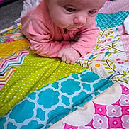 Lennon is registered to the contest to win money with this photo: baby, infant, child, pink_clothing, patchwork_quilt, colorful, patterned, fabric, lying_down, curious, indoor, closeup, hands, face, head, playtime, soft, cute, young, exploration