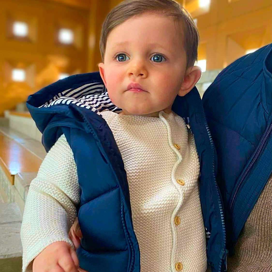 Aubin a rejoint le concours — aidez-le/la à gagner de superbes lots ! adult, baby, blue_eyes, casual, child, cute, expression, face, fashion, hand, holding, indoor, knit_sweater, person, portrait, puffer_vest, staircase, toddler, warm_clothing, wooden_wall