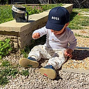Liam participe au concours pour gagner de l'argent avec cette photo : cap, casual, child, flowers, garden, grass, gravel, greenery, hat, mug, outdoor, pants, playful, shadow, sitting, stone_wall, stones, sunlight, toddler, white_clothing