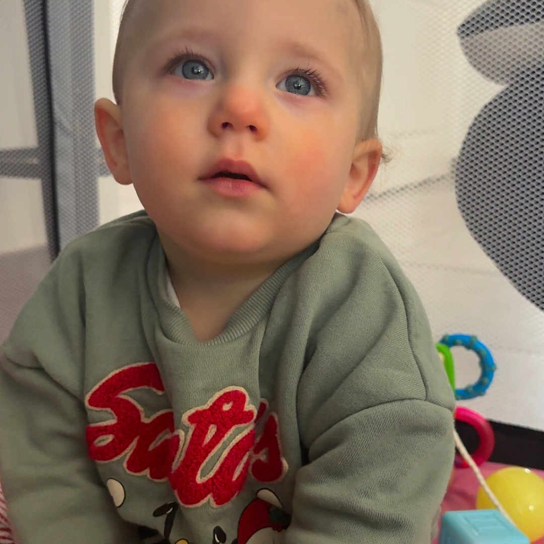 Liam a rejoint le concours — aidez-le/la à gagner de superbes lots ! toddler, child, baby, blue_eyes, sweater, striped_pants, pink_blanket, toys, indoor, mesh_barrier, curious, sitting, playtime, cute, portrait, young_child, face, hands, expression, soft_lighting